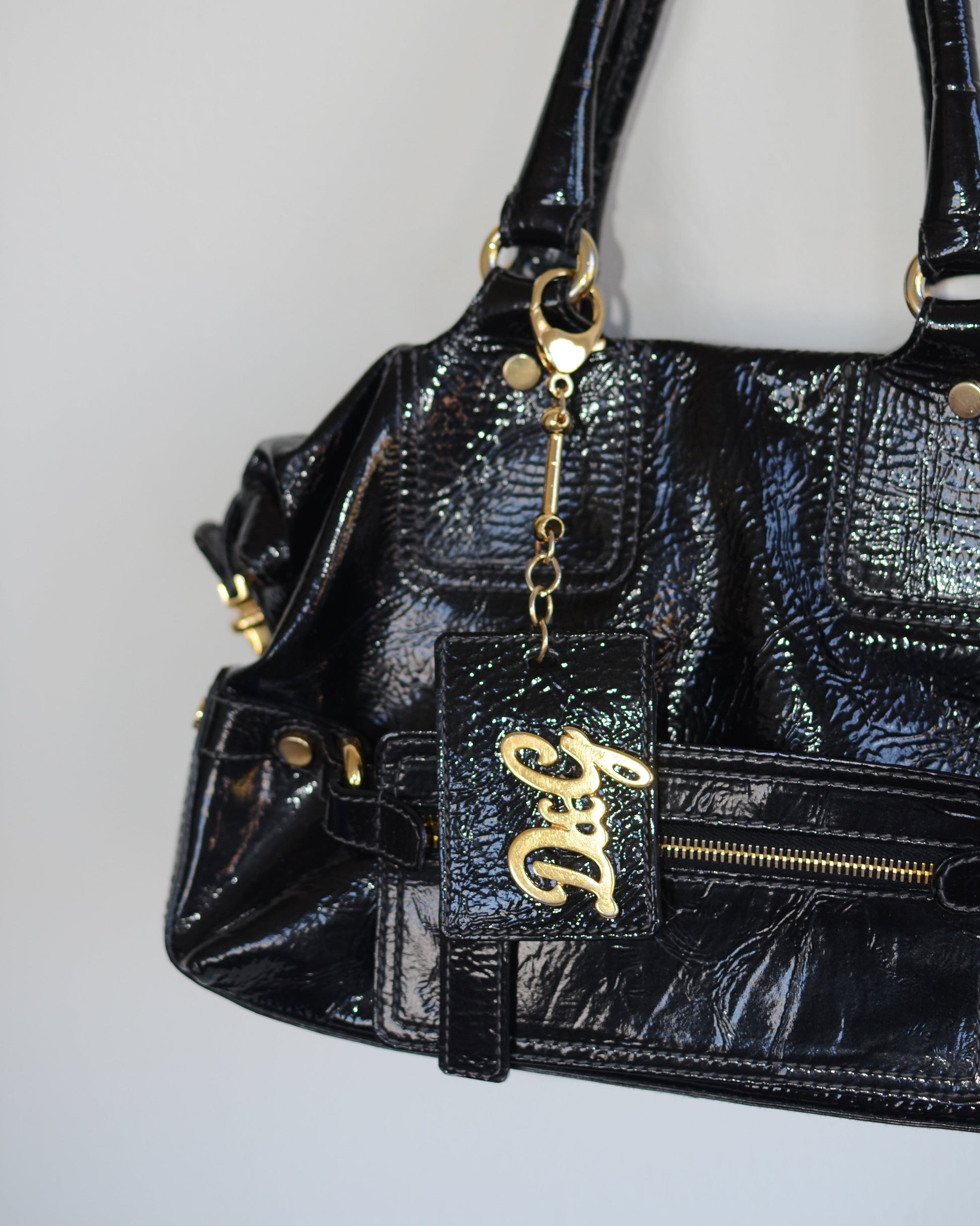 Close up of Dolce & Gabbana Black patent leather handbag with gold accents and hardware, with gold D&G logo charm on a white background