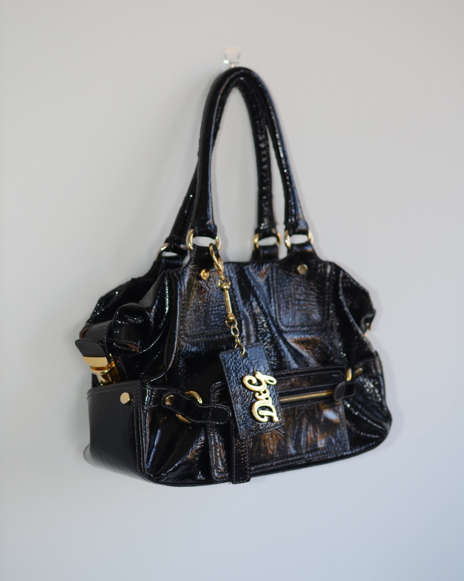 Side view of Dolce & Gabbana Black patent leather handbag with gold accents and hardware, with gold D&G logo charm on a white background
