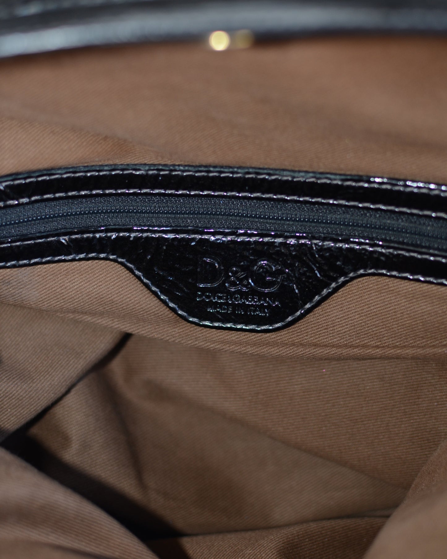 Dolce & Gabbana logo inside lining Black patent leather handbag with gold accents and hardware, with gold D&G logo charm on a white background