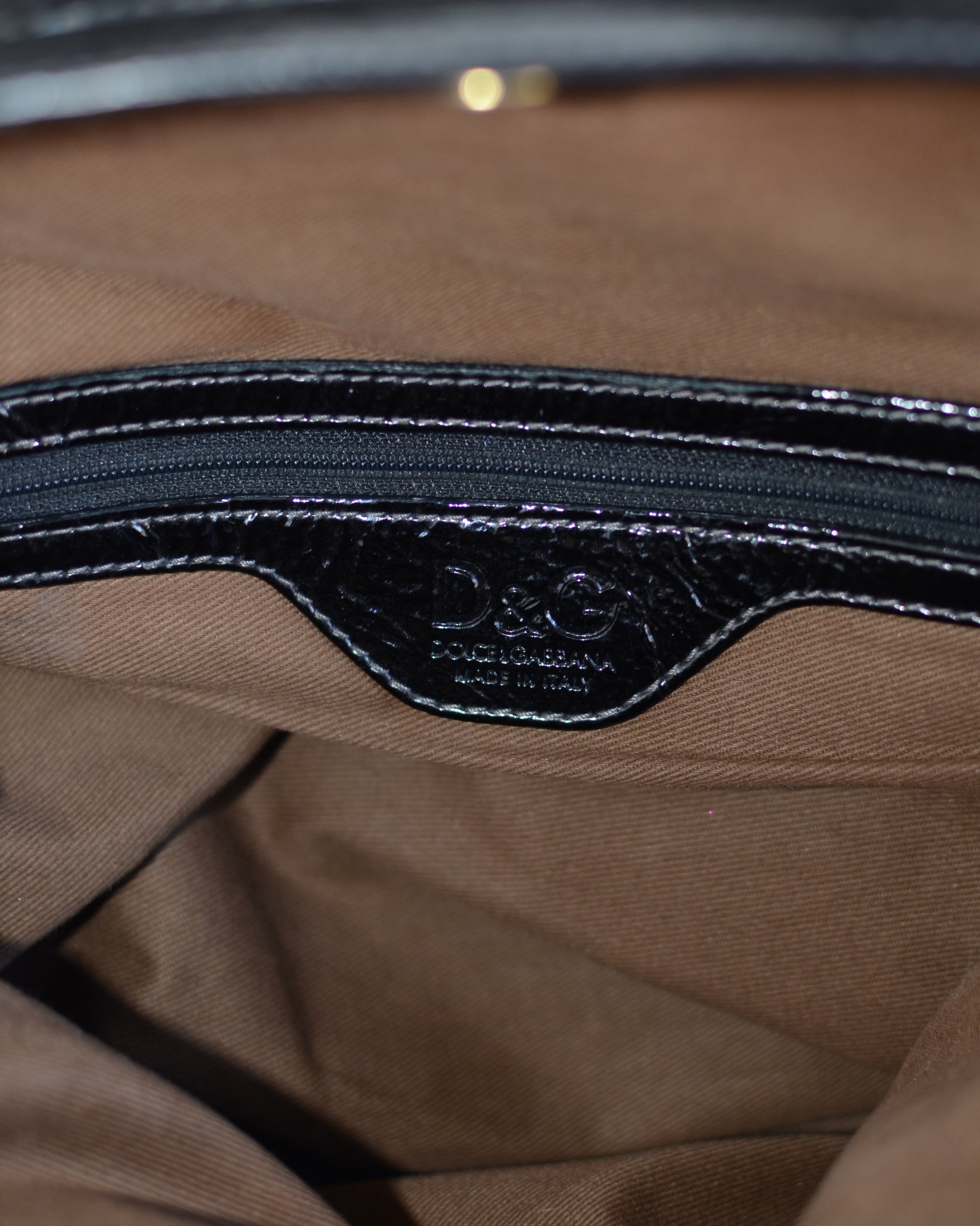 Dolce & Gabbana logo inside lining Black patent leather handbag with gold accents and hardware, with gold D&G logo charm on a white background