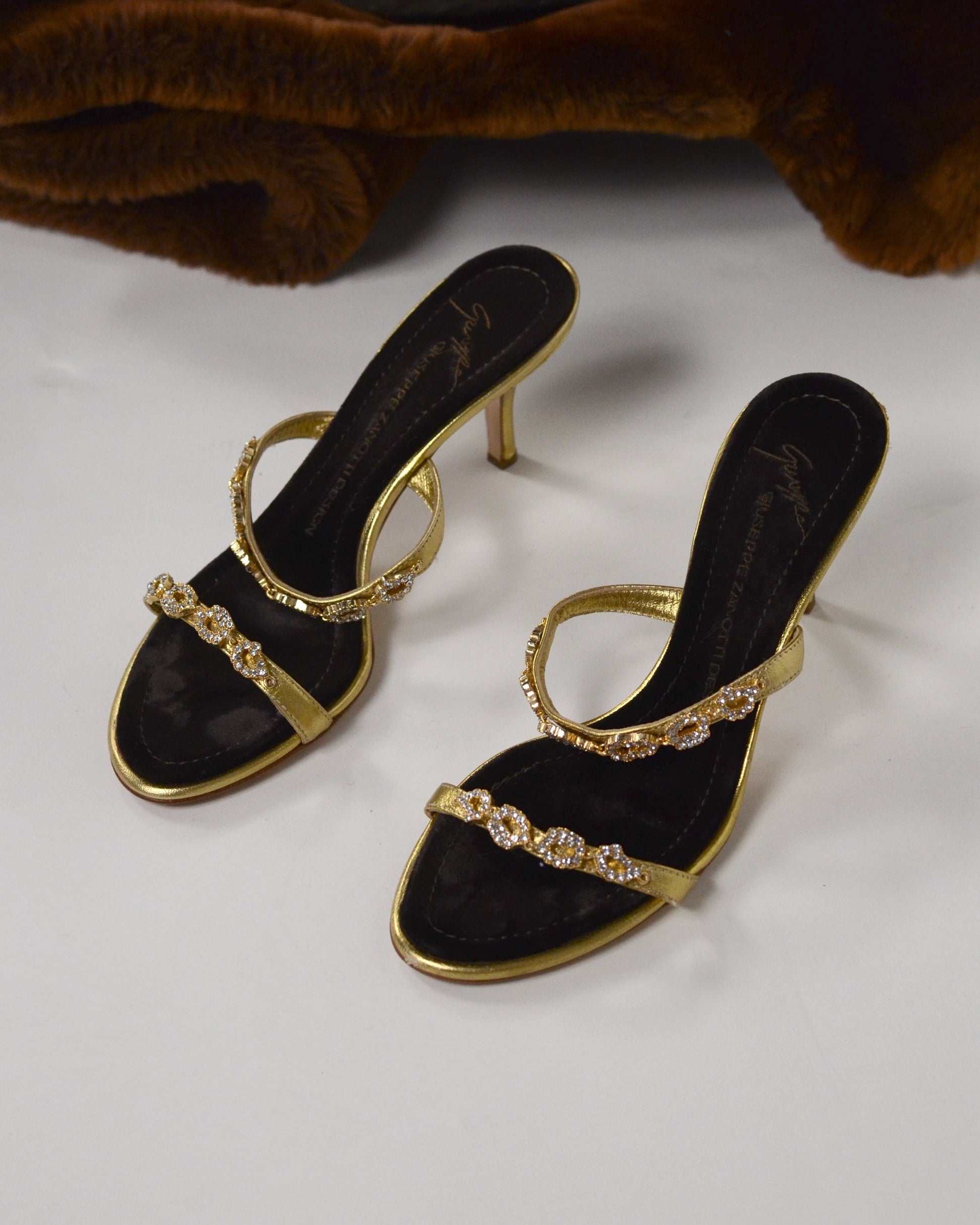 Gold high-heeled  Giuseppe Zanotti sandals with gold straps with  embellishments and brown velvet sole on a white surface.