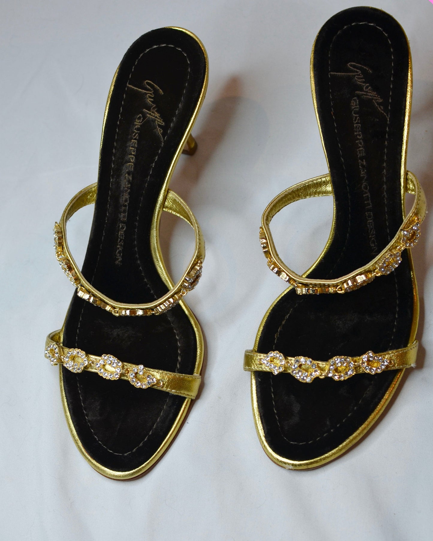 Gold high-heeled  Giuseppe Zanotti sandals with gold straps with  embellishments and brown velvet sole on a white surface.