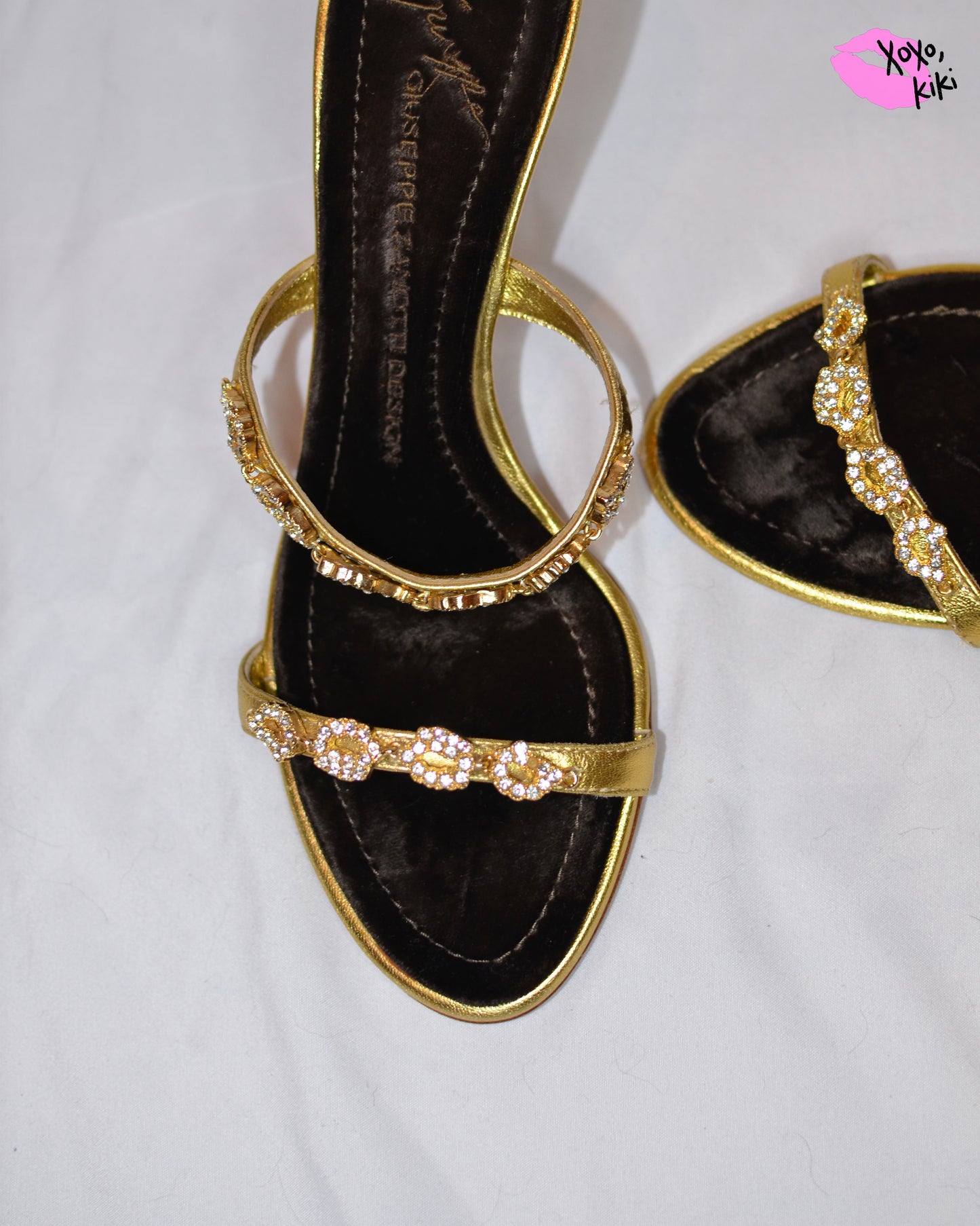 Gold high-heeled  Giuseppe Zanotti sandals, close up on gold straps with  embellishments and brown velvet sole on a white surface.