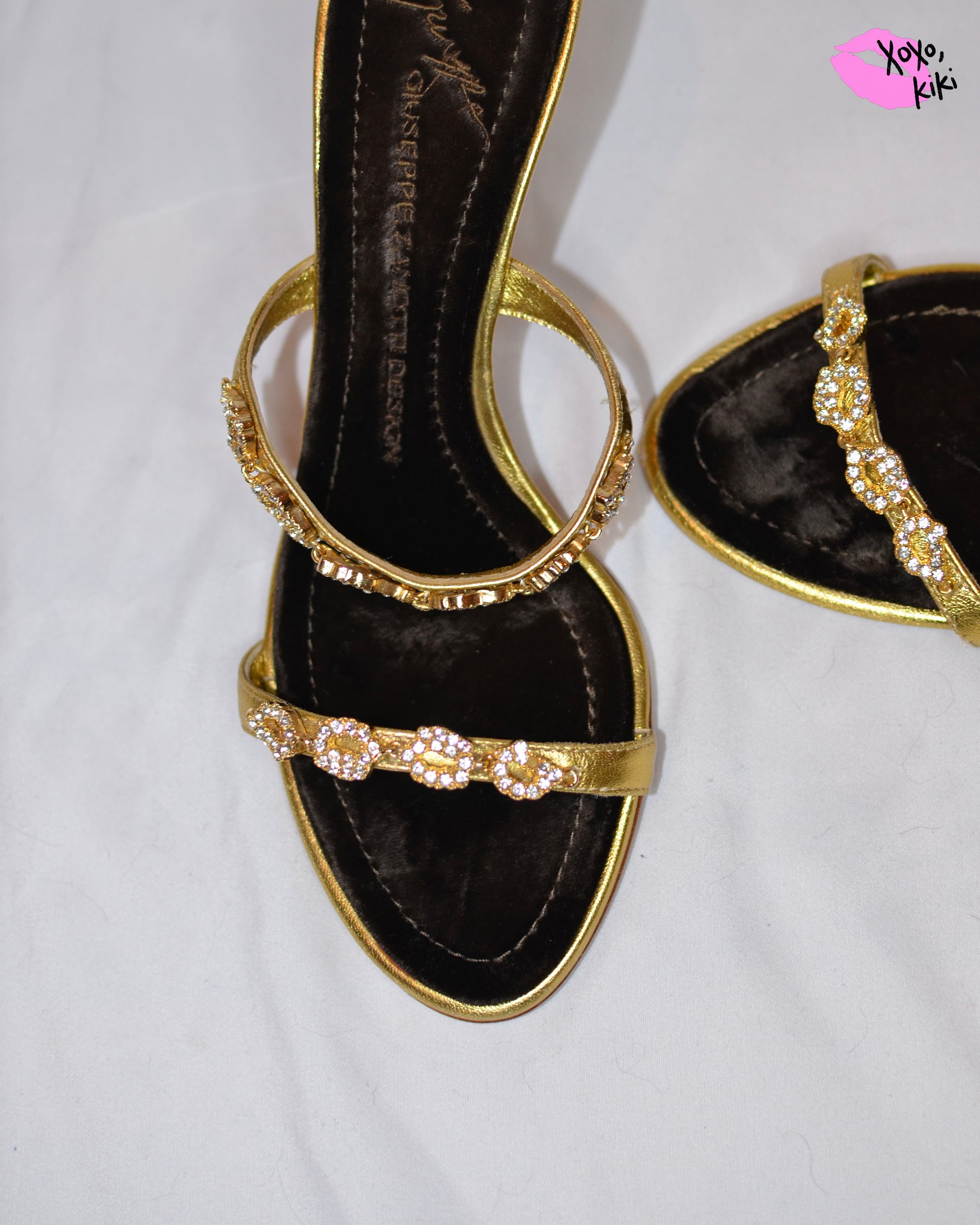 Gold high-heeled  Giuseppe Zanotti sandals, close up on gold straps with  embellishments and brown velvet sole on a white surface.